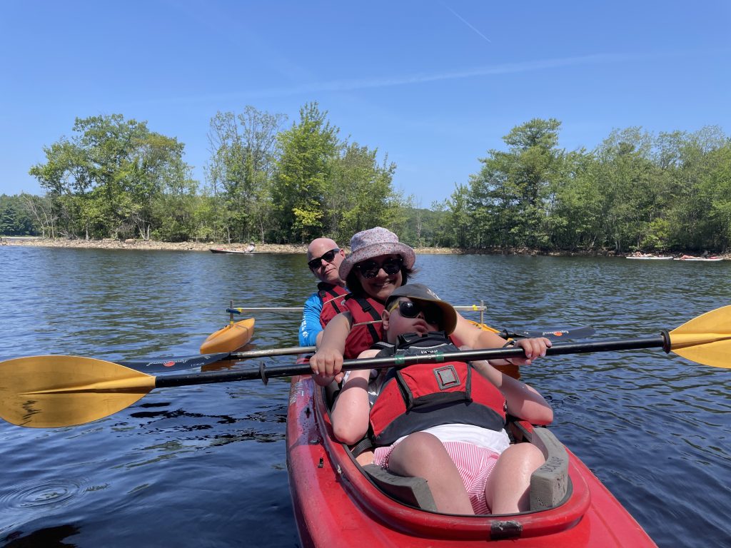 A dad, mom, and teenager sitting in a red kayak on a lake. All are wearing sunglasses, and Mom and Teen are holding a kayak paddle.