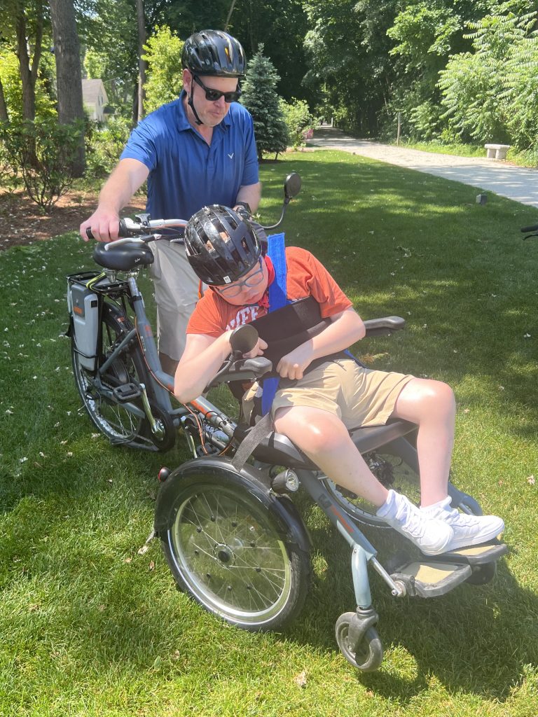 A man guides his teenage son in an adaptive bicycle. Both are wearing sunglasses, and they are about to enter a paved trail to ride.