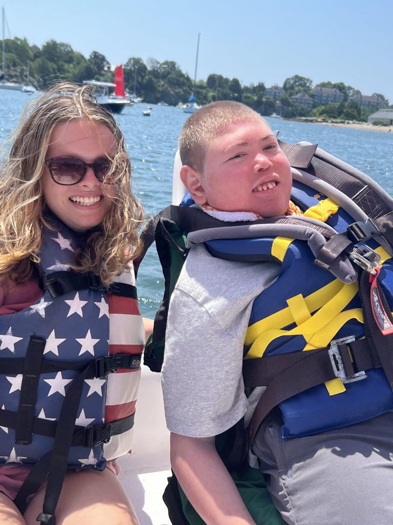 A teenage girl wearing an American flag life vest is sitting next to a teenage boy wearing a blue life jacket. They are on a sailboat, with the water behind them, and they are smiling.
