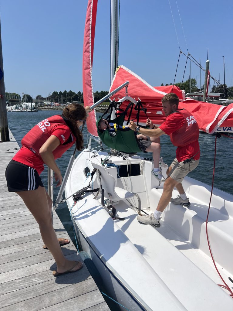 A young man and a young woman wearing red shirts with the words Sail To Prevail are guiding a teenage boy in a Hoyer sling onto a seat on a sailboat docked at a pier. The seat is fitted with a five-point harness.